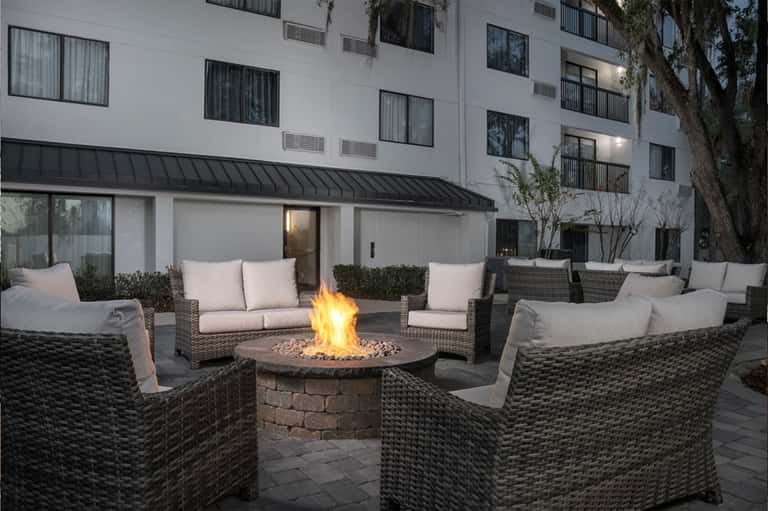 Outdoor fire pit lounge area with wicker seating and cushions in front of modern hotel building