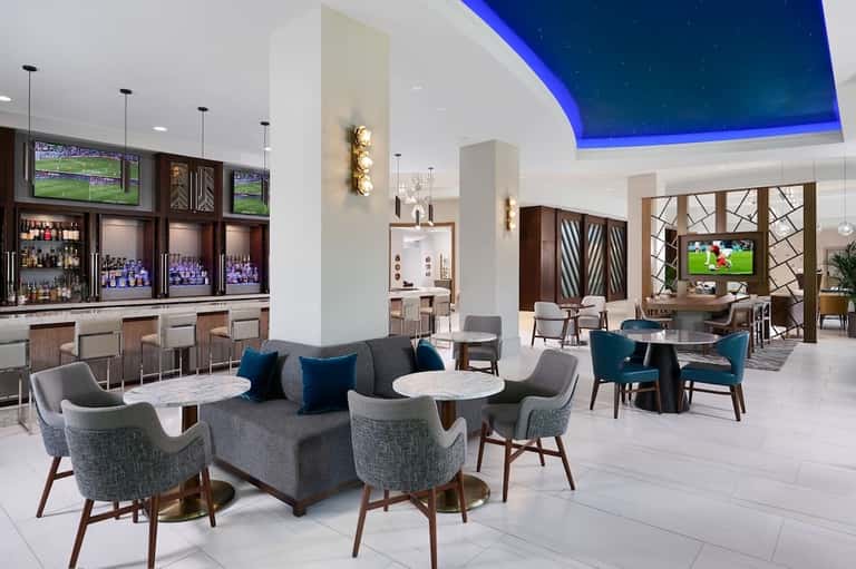 Modern hotel lobby lounge with blue-lit ceiling, contemporary furniture, bar, and flat-screen TVs