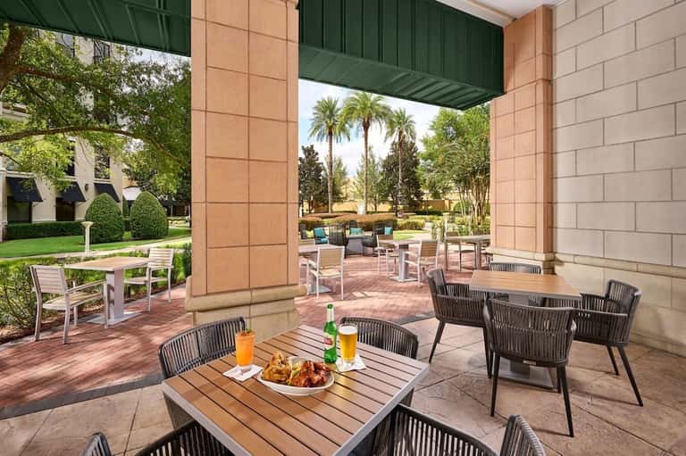 Covered outdoor patio with wooden tables, rope chairs, beverages, and view of tropical landscaping