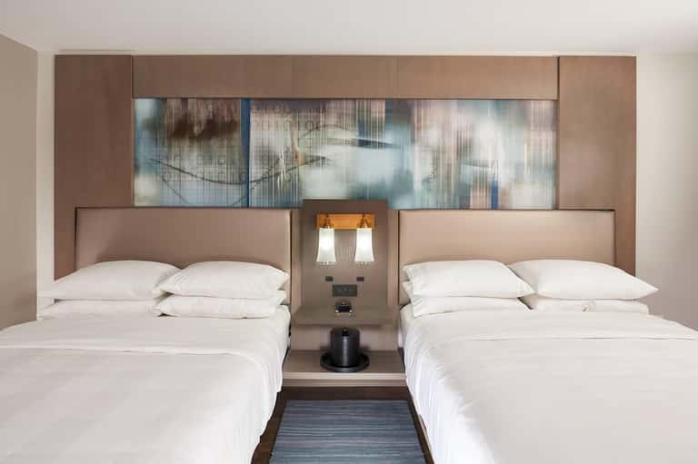 Hotel room interior with two queen beds, taupe upholstered headboards, and modern lighting fixtures