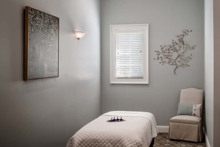 Minimalist spa treatment room with massage table, neutral walls, window with shutters, and botanical wall art