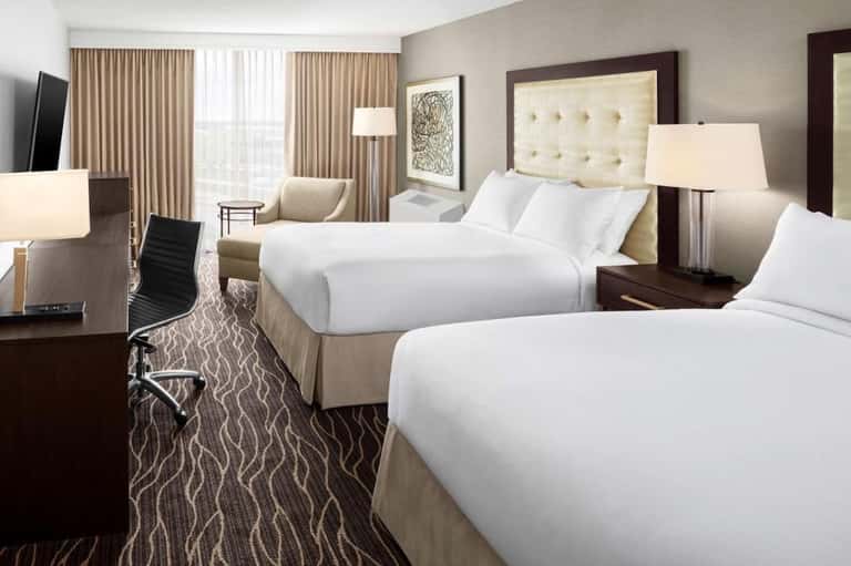 Hotel bedroom with two queen beds, beige headboards, dark wood furniture, and large windows