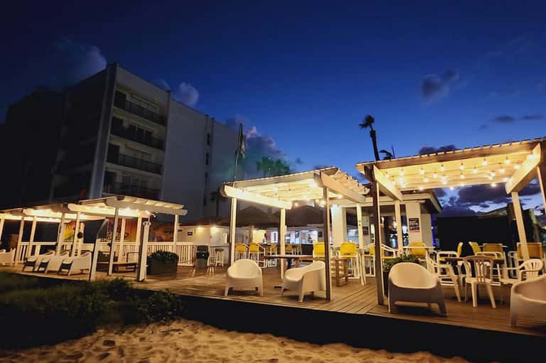 Illuminated beachfront deck pavilions at twilight with lounge seating, wooden pergolas, and sand