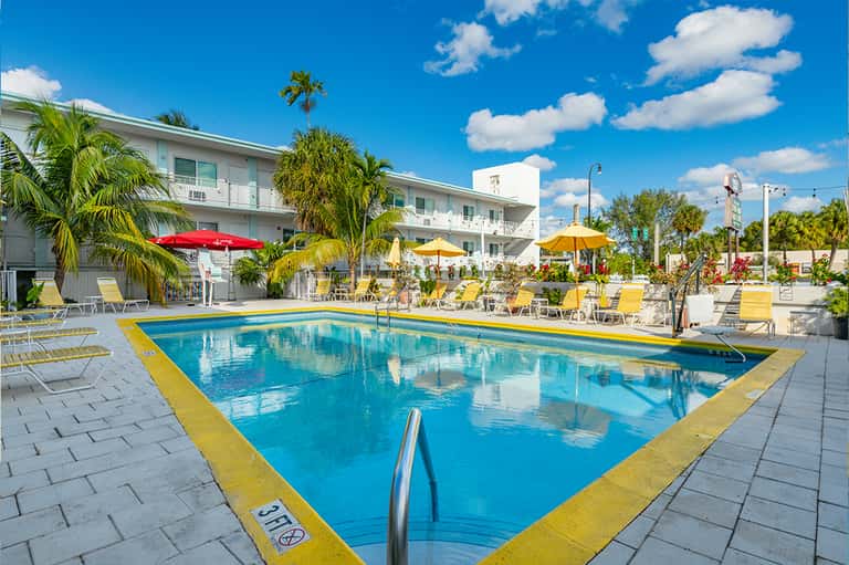 Resort swimming pool with yellow umbrellas, palm trees, and multi-story white building