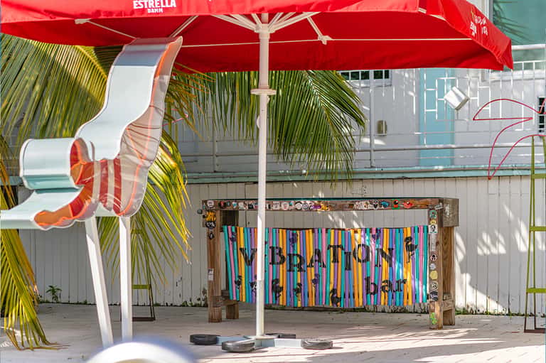 Colorful beachside art installation under red Estrella umbrella with palm trees and sandy area