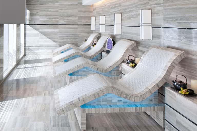Indoor spa facility with curved loungers around heated pools in neutral tones and natural light