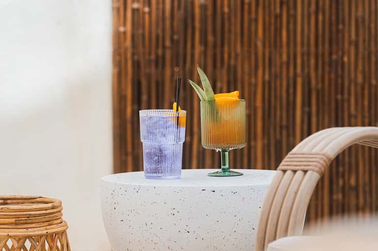 Two colorful cocktails on concrete table with woven chair and brown curtain backdrop