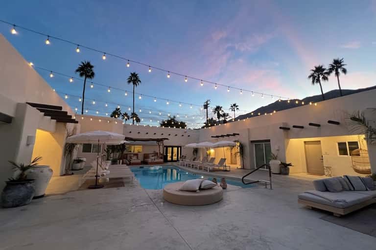 Resort courtyard at dusk with illuminated pool, fire pit, loungers, string lights, and palm trees