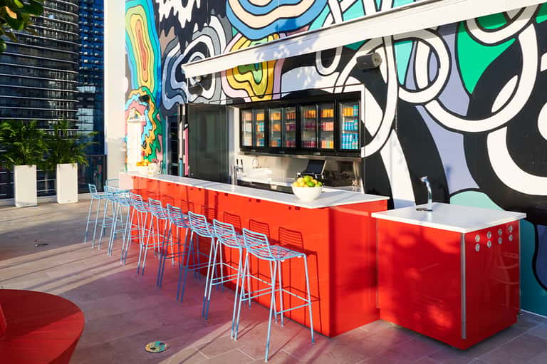 Colorful outdoor bar with vibrant street art mural, red counter seating, and beverage coolers