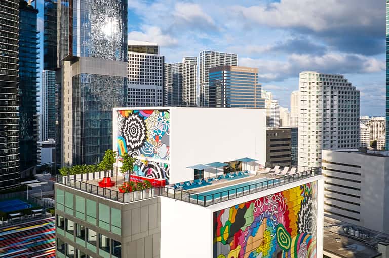 Urban rooftop pool deck with colorful mural art, lounge chairs, and city skyline views among high-rise buildings