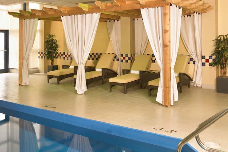 Spa relaxation area with wooden pergola, white curtains, yellow cushioned lounge chairs, and decorative tile border