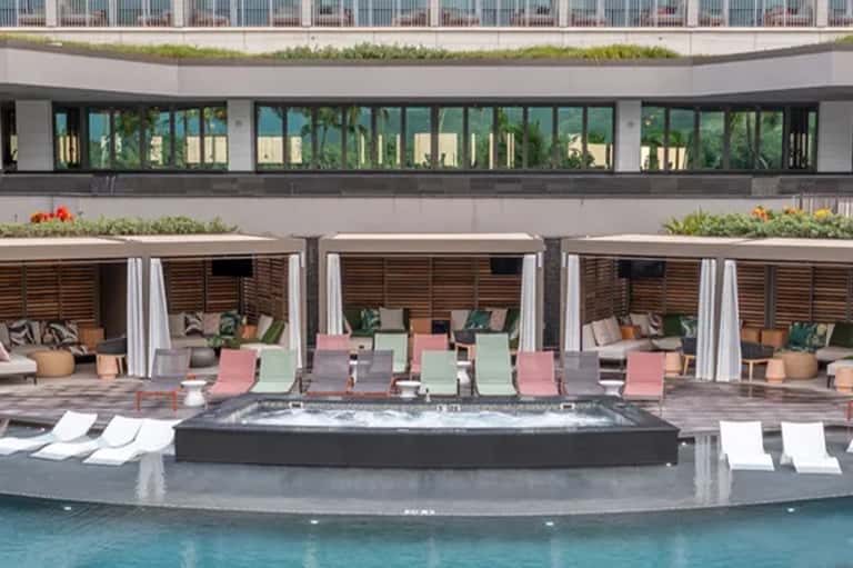 Resort pool deck with multi-colored loungers, cabanas with curtains, hot tub, and modern building