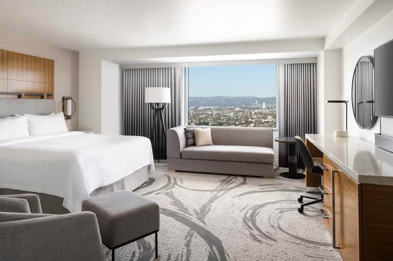Upscale hotel bedroom with king bed, gray sofa, floor-to-ceiling windows overlooking cityscape