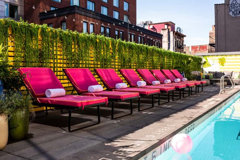 Urban rooftop pool deck with hot pink loungers, ivy wall, city buildings, and turquoise pool