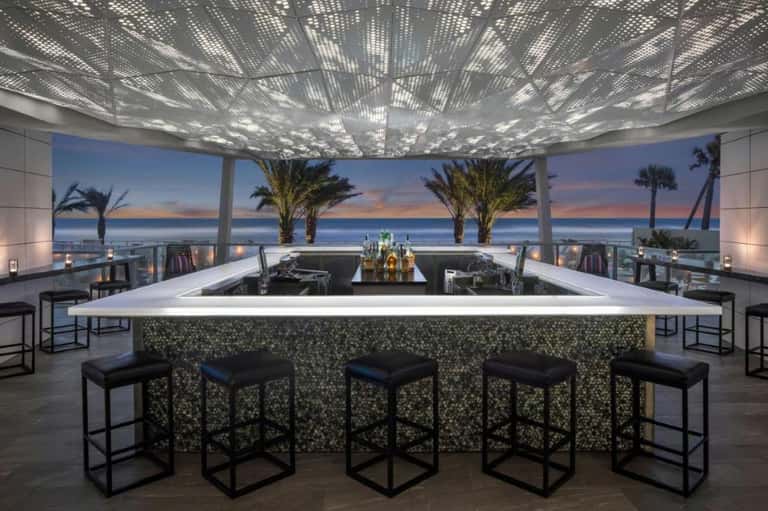 Rooftop bar with panoramic ocean and sunset views, perforated ceiling, modern counter seating, and palm trees