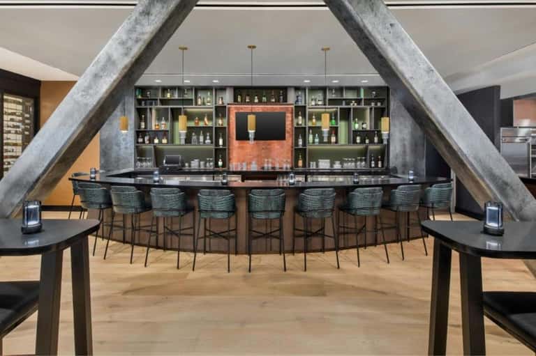 Contemporary hotel bar with curved seating, large concrete support beams, extensive bottle display, and ambient lighting