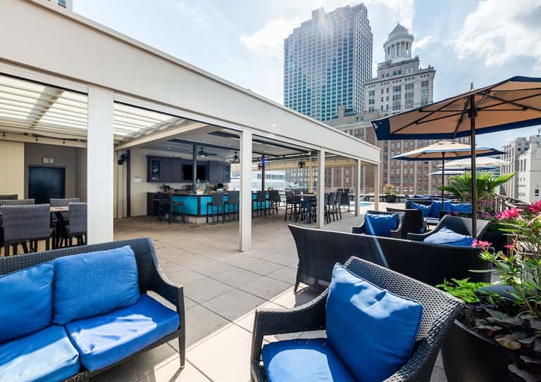 Rooftop terrace with modern lounge chairs featuring blue cushions under extended pergola with city skyline views and blue bar