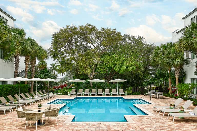 Resort-style pool area with lounge chairs, white umbrellas, palm trees, and manicured landscaping