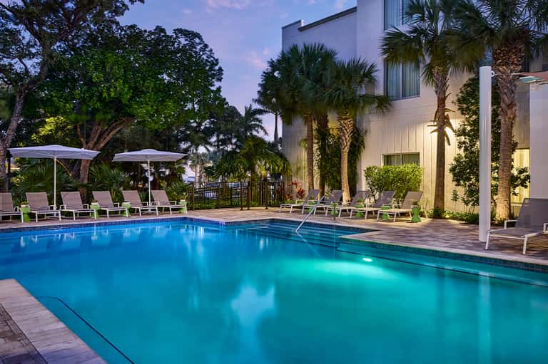 Illuminated resort pool at dusk with lounge chairs, umbrellas, palm trees, and modern white building