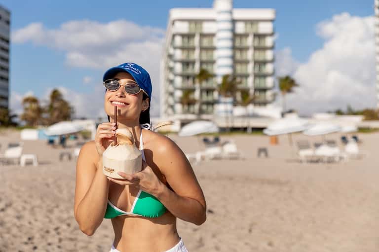Woman on beach holding coconut drink with high-rise resort buildings in background