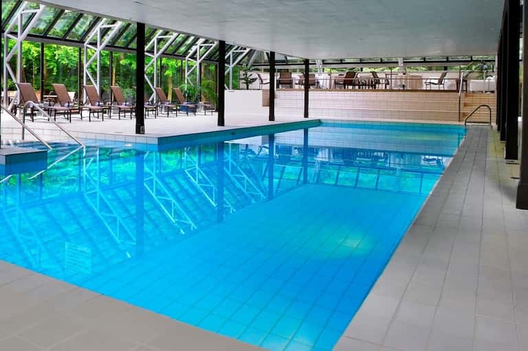 Indoor pool facility with glass ceiling, turquoise water, white lane markers, metal structural framework, and lounge area.