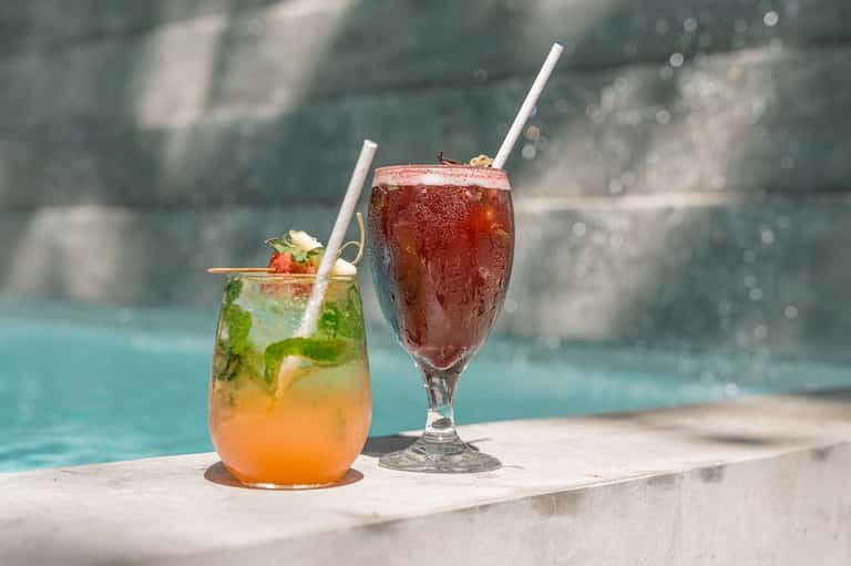 Two colorful cocktails on pool deck ledge with turquoise water and stone wall backdrop