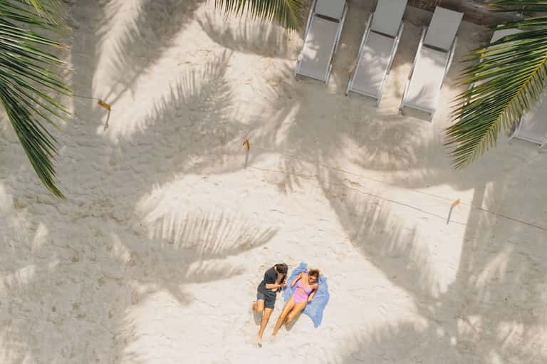 Overhead view of two guests relaxing on white sand beach between palm fronds
