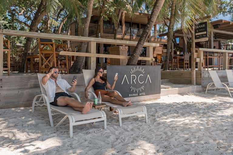 Beachfront lounge area with palm trees, ARCA Boutique Hotel signage, and guests relaxing on white loungers