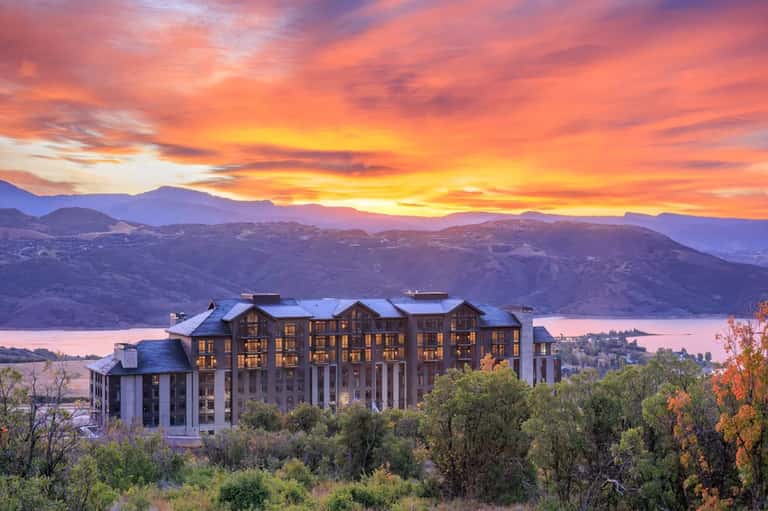 Upscale resort building at sunset with illuminated windows overlooking a lake and mountains at dusk