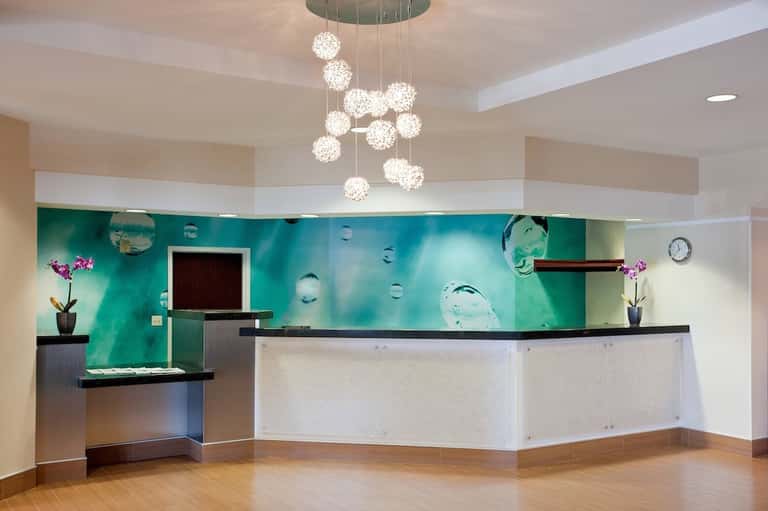 Modern hotel lobby with turquoise water-themed mural, hanging crystal chandelier, white desk, and wooden accents