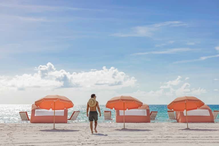 Beach scene with surfer walking toward orange and white beach umbrellas on sandy shore with ocean and clouds.
