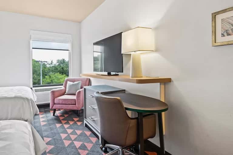 Hotel room with two beds, desk area, pink accent chair, and balcony with natural light