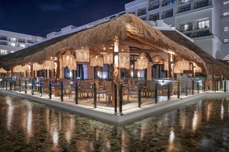 Illuminated thatched-roof beachfront restaurant with wooden chairs overlooking water