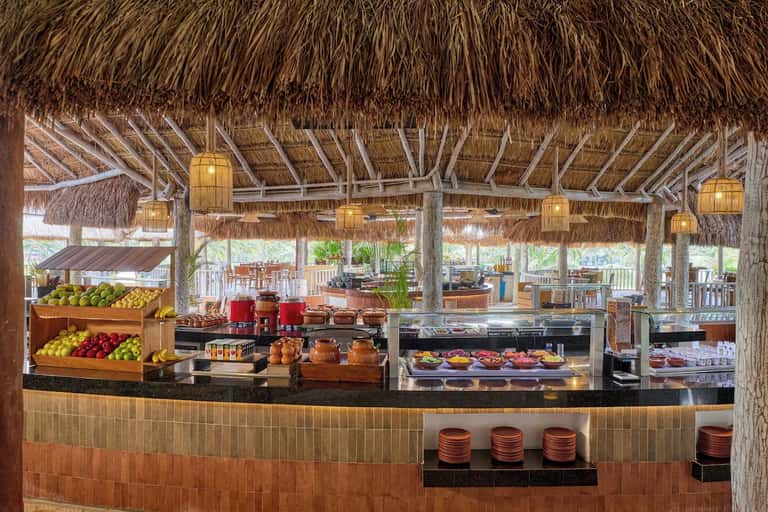 Outdoor beachfront bar with thatched roof, fresh fruit display, and ocean backdrop