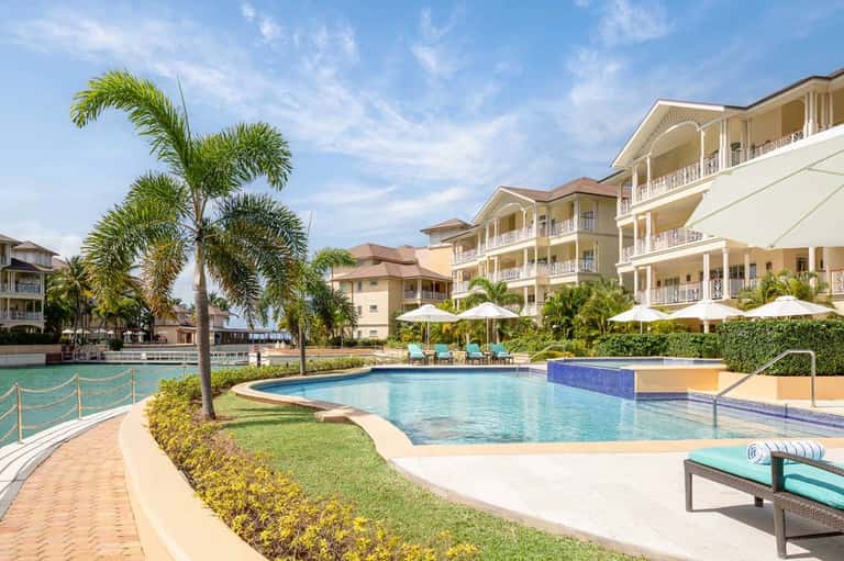 Resort exterior with multi-story buildings, pool, palm trees, white umbrellas, and manicured lawn