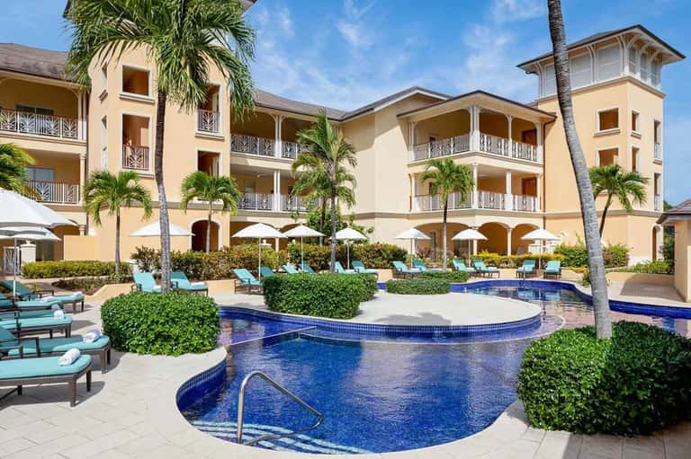 Resort courtyard with curved swimming pool, lounge chairs, white umbrellas, and multi-story yellow building