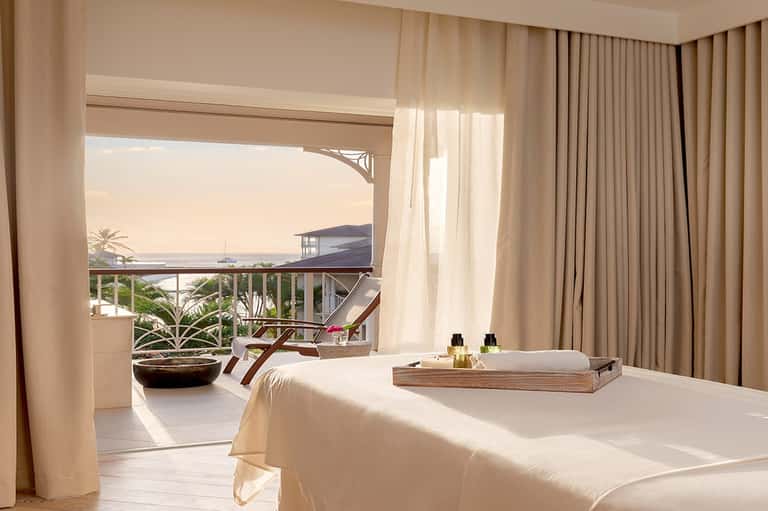 Luxury spa treatment room with massage bed, ocean and mountain views from balcony, cream curtains, and daybed seating