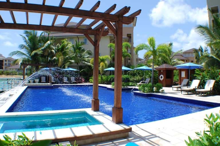 Resort swimming pool with wooden pergola, palm trees, blue umbrellas, and lounge chairs
