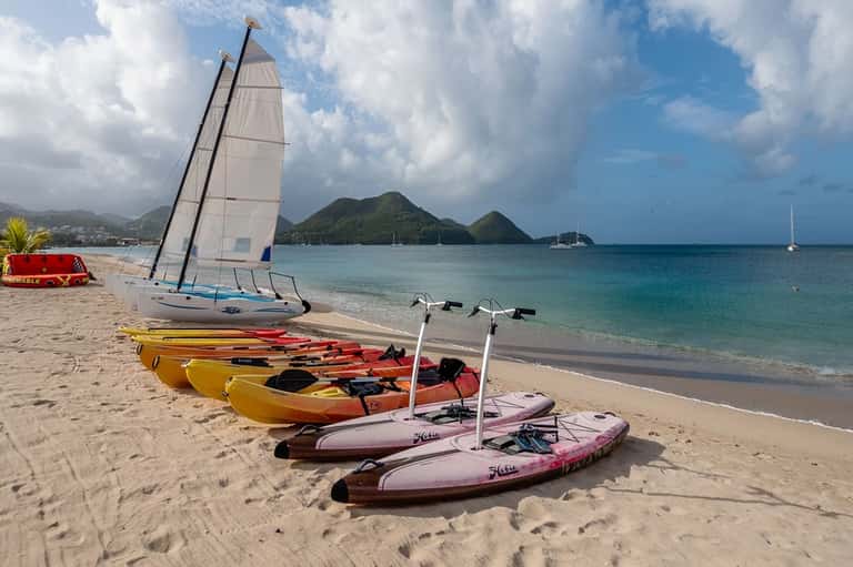 Beach scene with sailboat, colorful kayaks, and paddleboards on sand near tropical bay