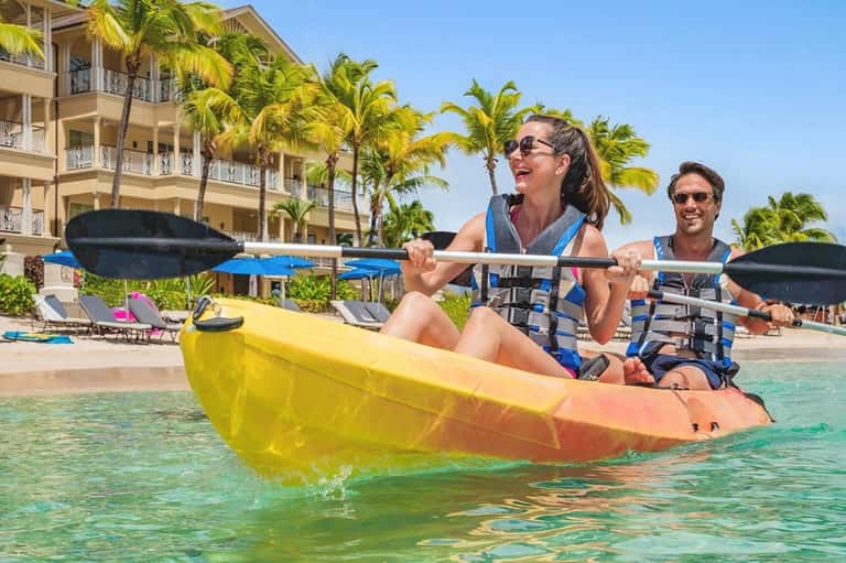 Couple kayaking in turquoise waters near resort buildings with palm trees in tropical setting