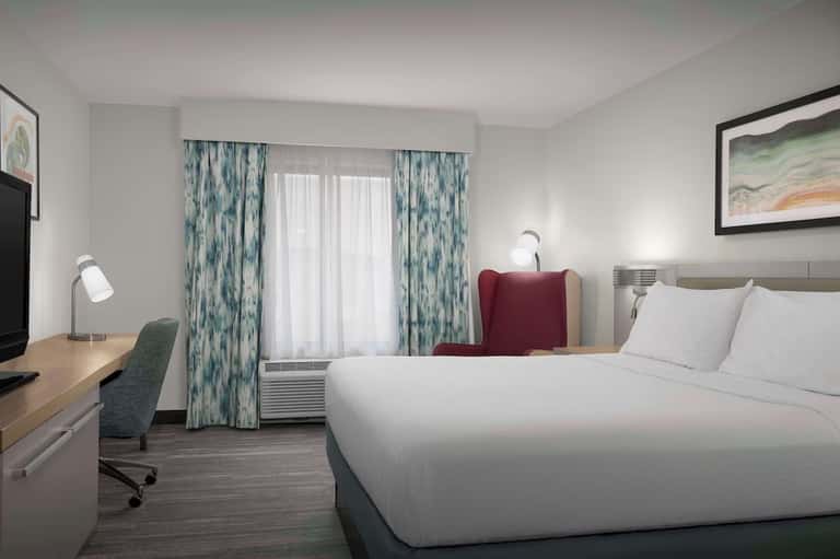 Modern hotel bedroom with queen bed, desk, patterned curtains, and flat-screen TV