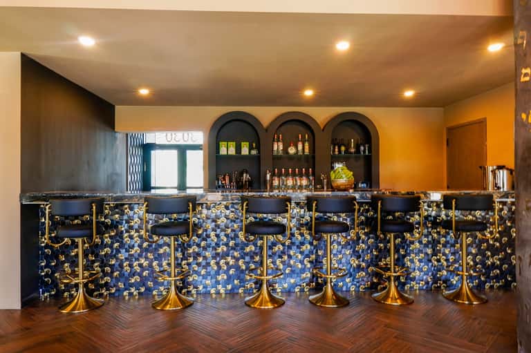Upscale bar with arched alcoves, blue-patterned backsplash, gold-trimmed chairs, and warm lighting.