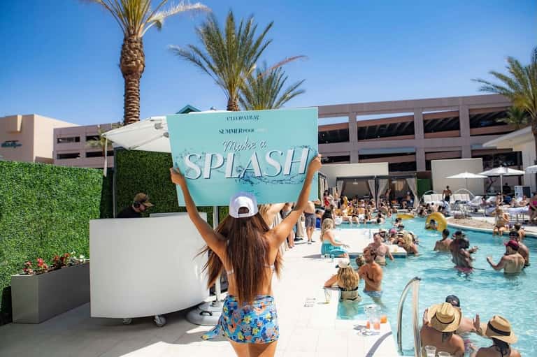 Woman holding turquoise splash sign at busy pool party with crowded swimming pool and palm trees