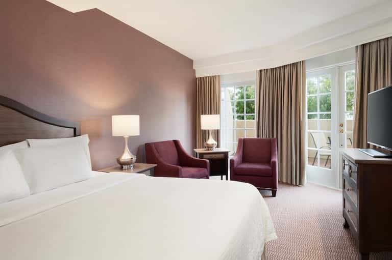 Hotel bedroom with queen bed, burgundy accent chairs, and balcony access
