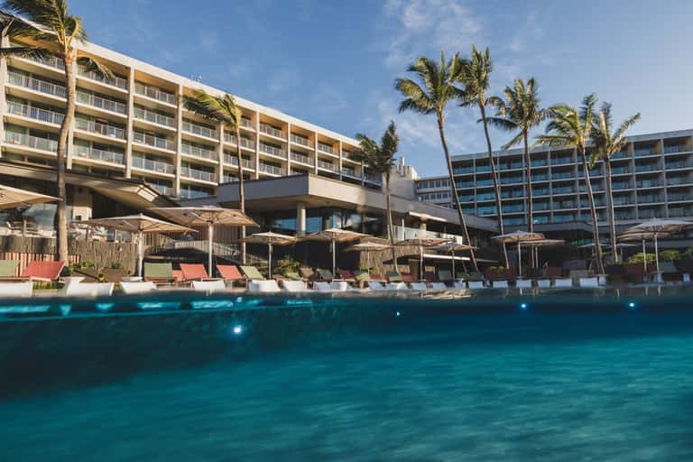 Beachfront resort with multi-story buildings, palm trees, pool deck with lounge chairs