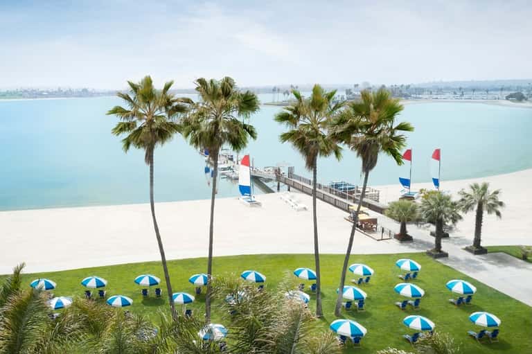 Aerial view of sandy beach with palm trees, wooden pier, blue and white umbrellas, and calm turquoise water