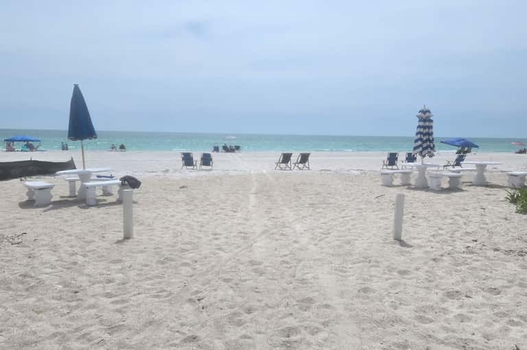 Beach scene with white picnic tables, black umbrellas, lounge chairs, and calm turquoise water