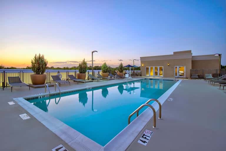 Twilight view of resort pool with lounge chairs, potted plants, waterfront, and lit building at dusk