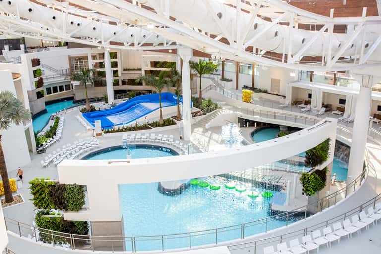 Indoor water park with multiple pools, white ceiling structure, blue water slides, and plants