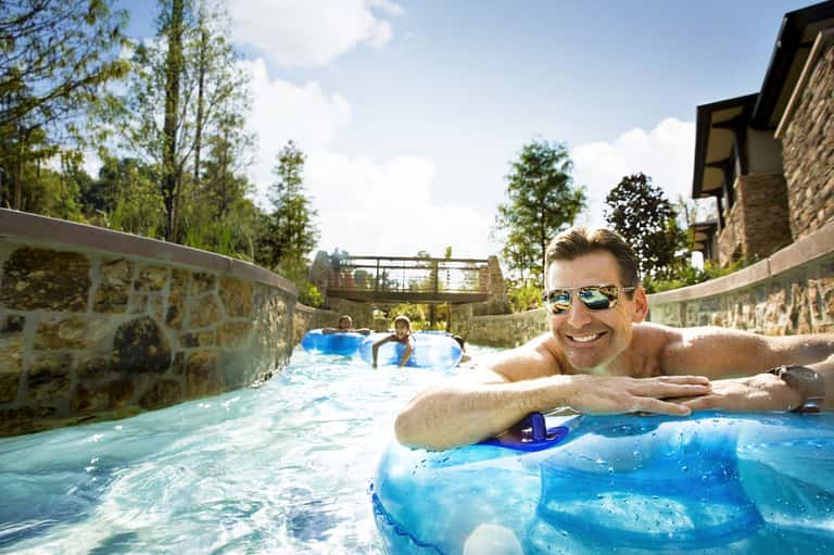 Man relaxing in a narrow resort pool with stone walls, surrounded by green trees and architectural features